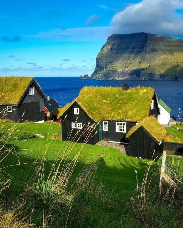 grass-roofed houses in the mikladalur village on the island of kalsoy and the cliffs of kunoy island at a distance, faroe islands.