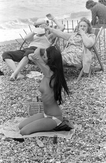 woman covering man's eyes with her knitting at sight of young woman taking off her top on the beach in france, 1974